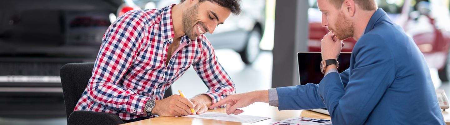 Man signing documents at desk with salesperson in dealership