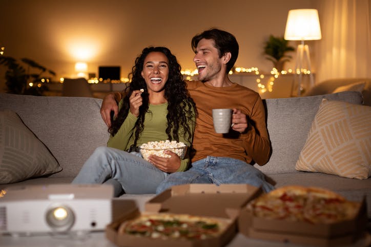 Cheerful Couple Using Home Cinema Projector Watching Comedy Movie Laughing, Eating Popcorn And Pizza Sitting On Sofa Indoors. Weekend Family Leisure, Entertainment And Fun