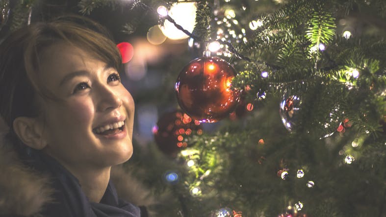 A young woman is standing by a big Christmas tree and smiling happily. The tree has many illumination lights and decorations. She is looking up the tree and fully smiling at night.