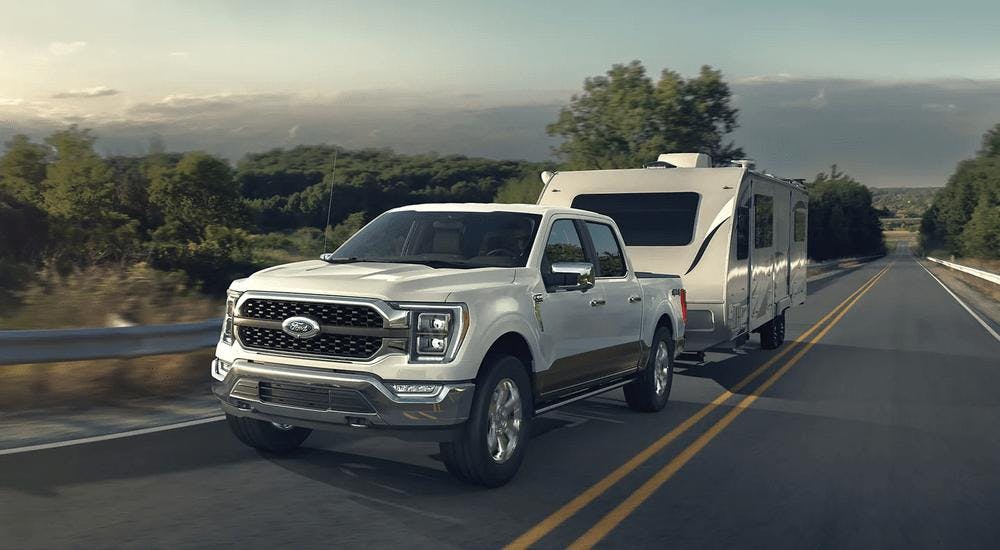 A white 2023 Ford F-150 is shown towing a camper on a highway.