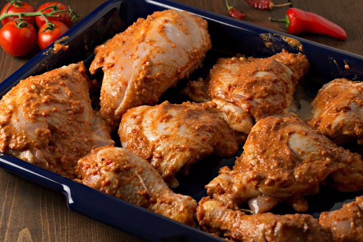 A high angle extreme close up shot of a blue metal tray with several pieces of chicken marinating in a spicy homemade African Harissa paste/sauce, the chicken will marinate for 24 Hours before being baked in the oven. The tray is framed by several ripe tomatoes and spicy chili peppers.