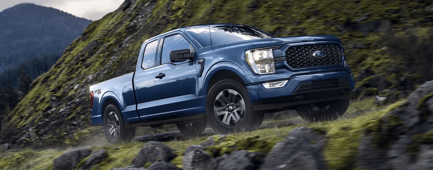 A blue 2023 Ford F-150 STX is shown driving up a mountain road.