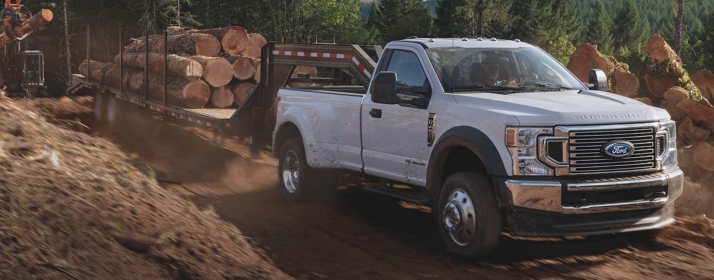 A white 2021 Ford F-450 Super Duty is shown towing a trailer full of logs after viewing used Ford trucks for sale.