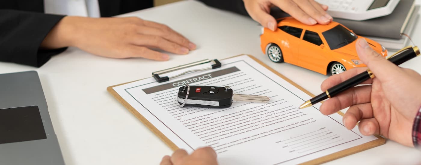 A person is shown filling out an auto loan form on a clipboard next to an orange toy car.