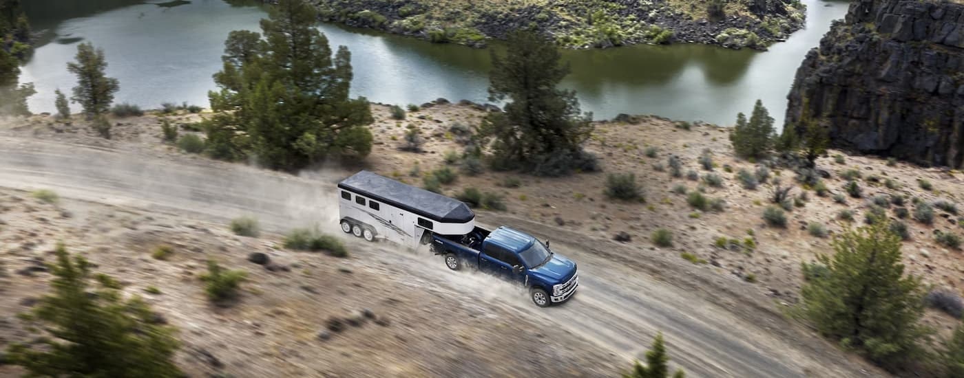 A blue 2023 Ford F-250 is shown towing a horse trailer on a dirt road.