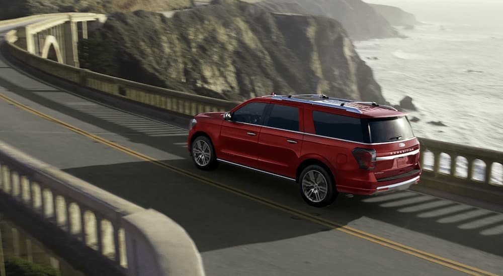 A red 2023 Ford Expedition Platinum is shown driving on a rocky highway next to a body of water.