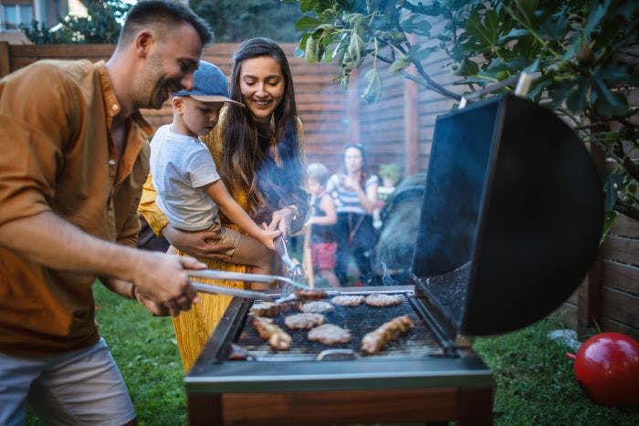 A man in an orange shirt stands beside his wife and son while he tends to the meat on a grill