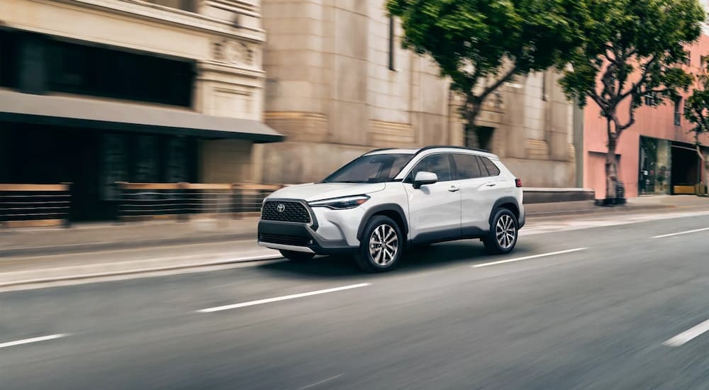 A white 2023 Toyota Corolla Cross XLE is shown driving on a city street after leaving a Toyota dealer in Westminster, VT.