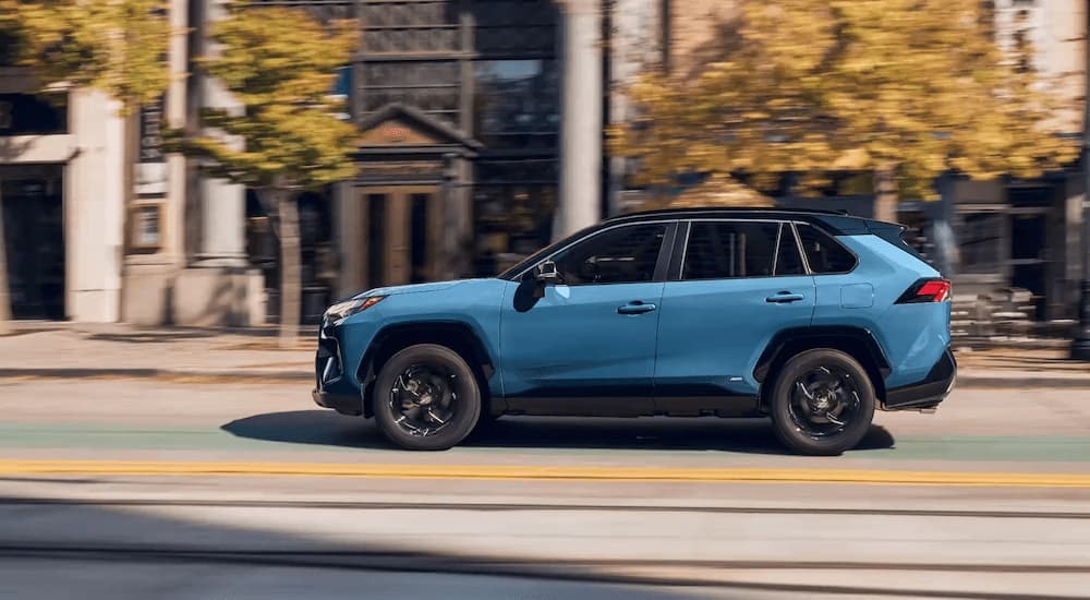 A blue 2023 Toyota RAV4 XSE Hybrid is shown from the side driving on a city street next to tall buildings.