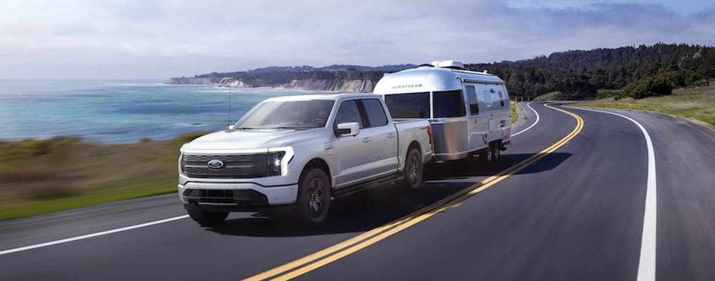 A white 2023 Ford F-150 Lightning is shown from the front at an angle while towing a camper.