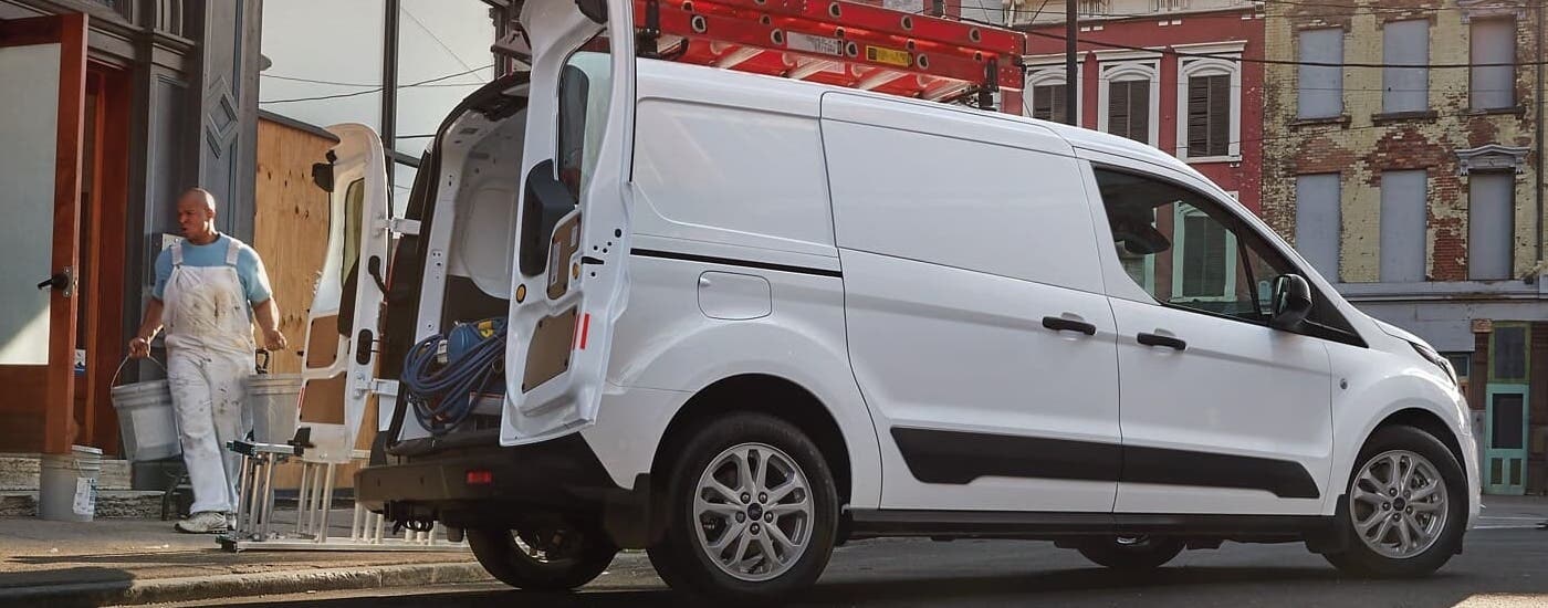 A white 2023 Ford Transit Connect is shown parked at a construction site with a ladder on the roof.