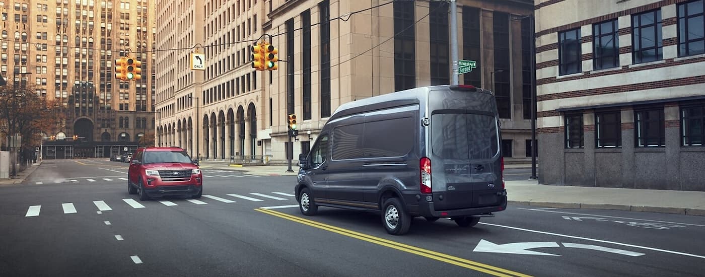 A grey 2023 Ford Transit is shown from a rear angle driving on a city street.