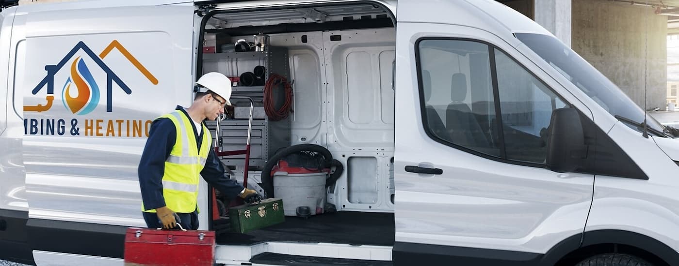 A plumber is shown next to a white 2023 Ford Transit with the cargo sliding door opened.