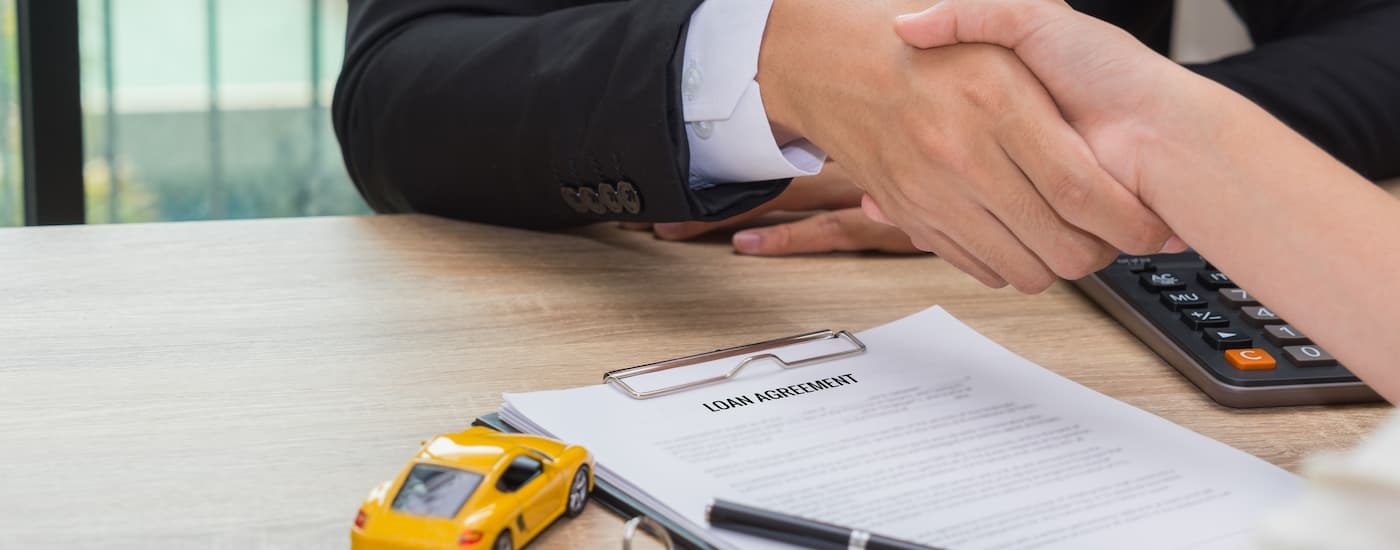 A salesman and customer are shown shaking hands over car loan paperwork.