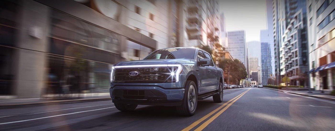 A blue 2024 Ford F-150 Lightning is shown driving on a city street.