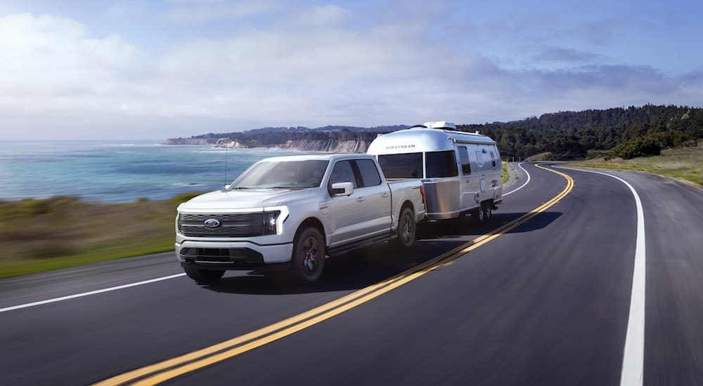 A white 2024 Ford F-150 Lightning is shown from the front while towing a camper.