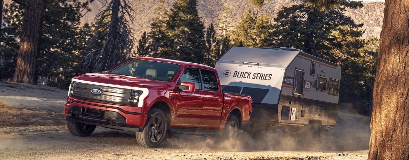 A red 2024 Ford F-150 Lightning is towing a camper on a dusty dirt road.