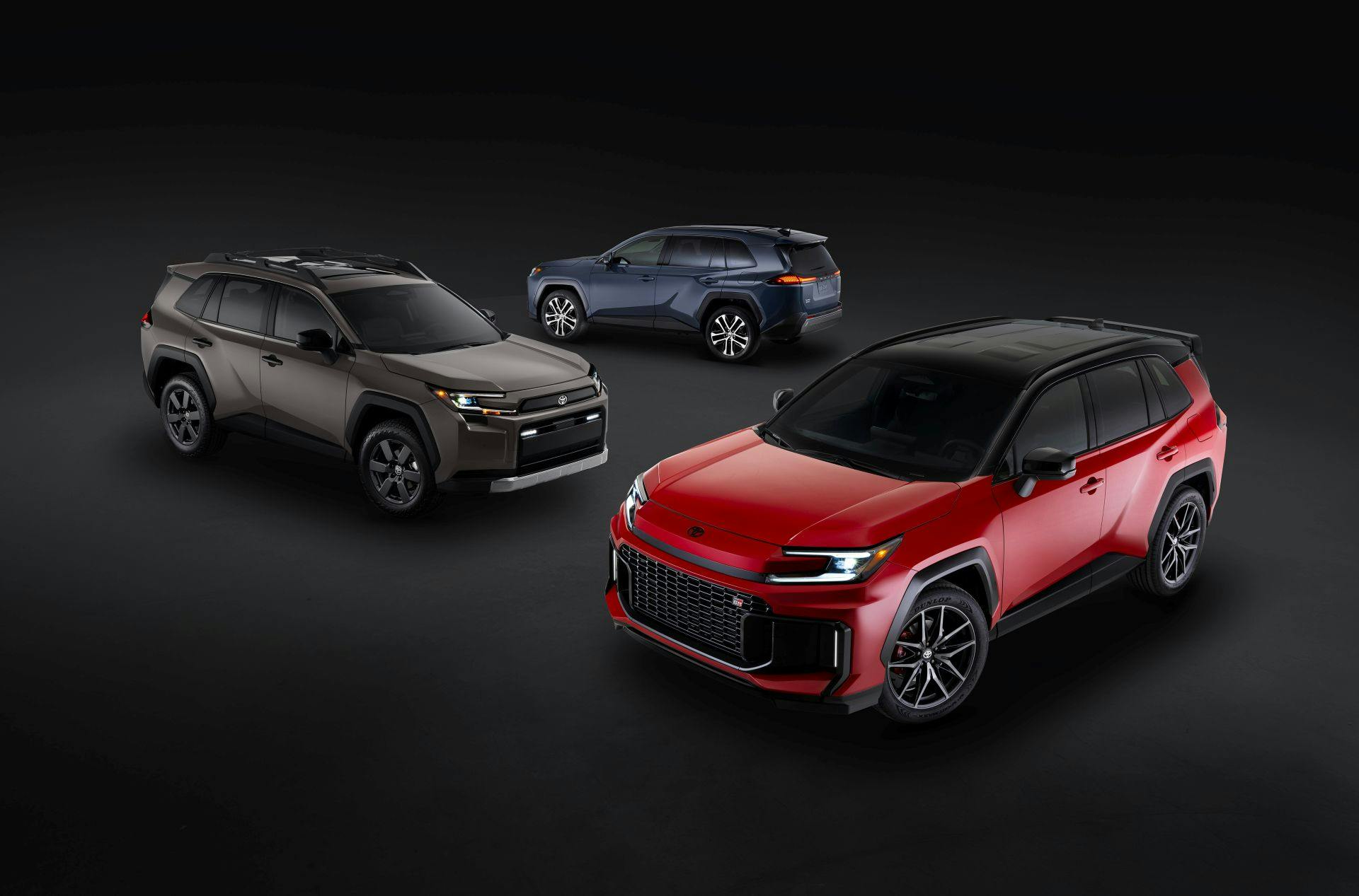Rendered image of three 2026 Toyota RAV 4 with a black background. Closest to furthest: Red GR Sport RAV4, Brown Woodland RAV4, and a Blue basic RAV4
