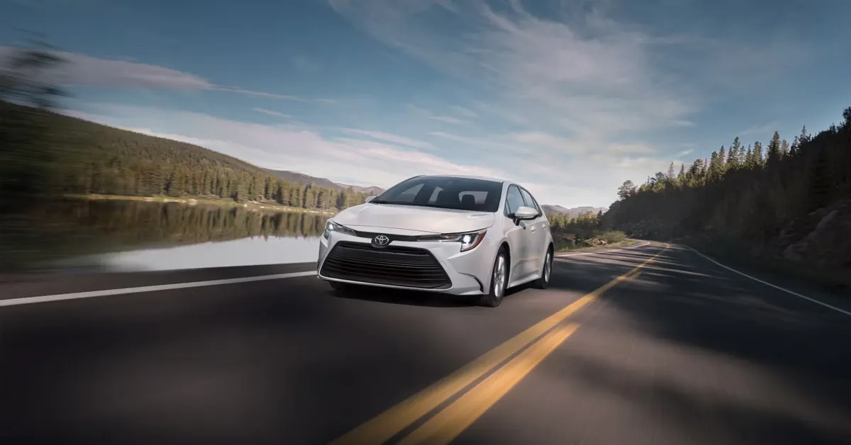 2026 Toyota Corolla driving along a scenic lakeside highway with forested mountains