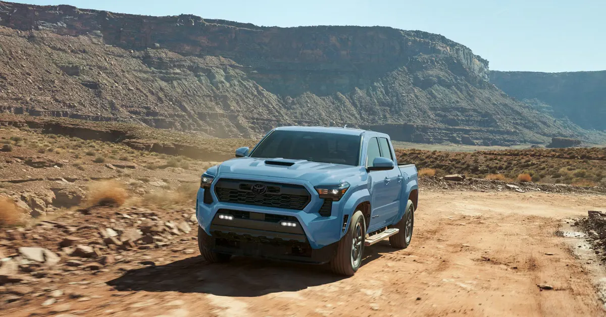 2026 Toyota Tacoma driving on a rugged desert trail with rocky canyon cliffs in the background, showcasing its off-road capability and bold exterior design.
