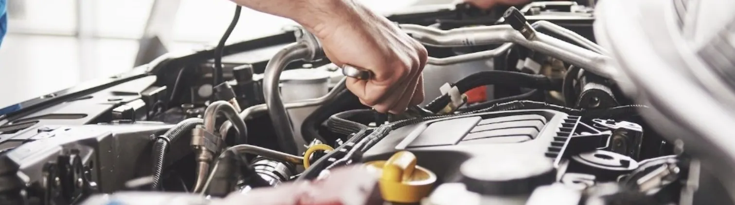 Automotive service technician working inside an open engine compartment while inspecting and servicing vehicle components.