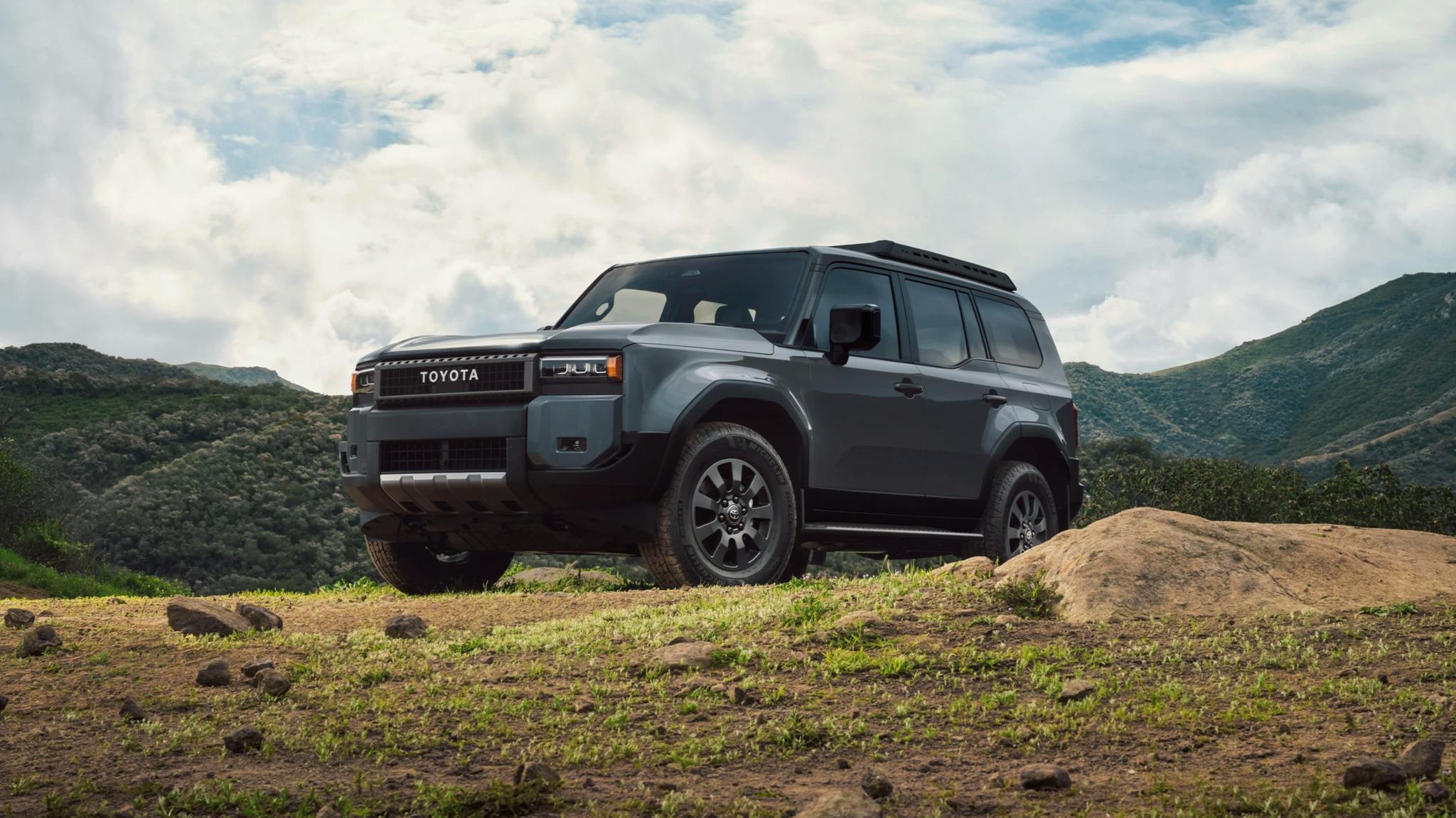 A Toyota Land Cruiser is parked majestically on a dirt road overlooking mountains in the background.