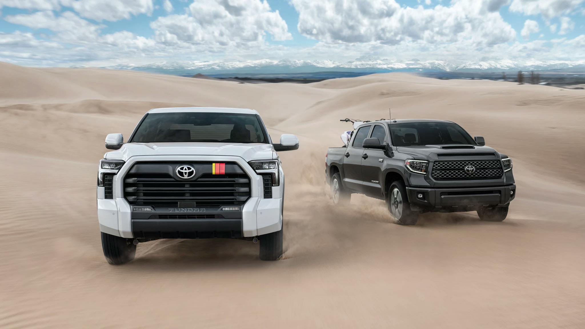 Two Toyota Tundra trucks driving on sand hills with mountains in the distant background.