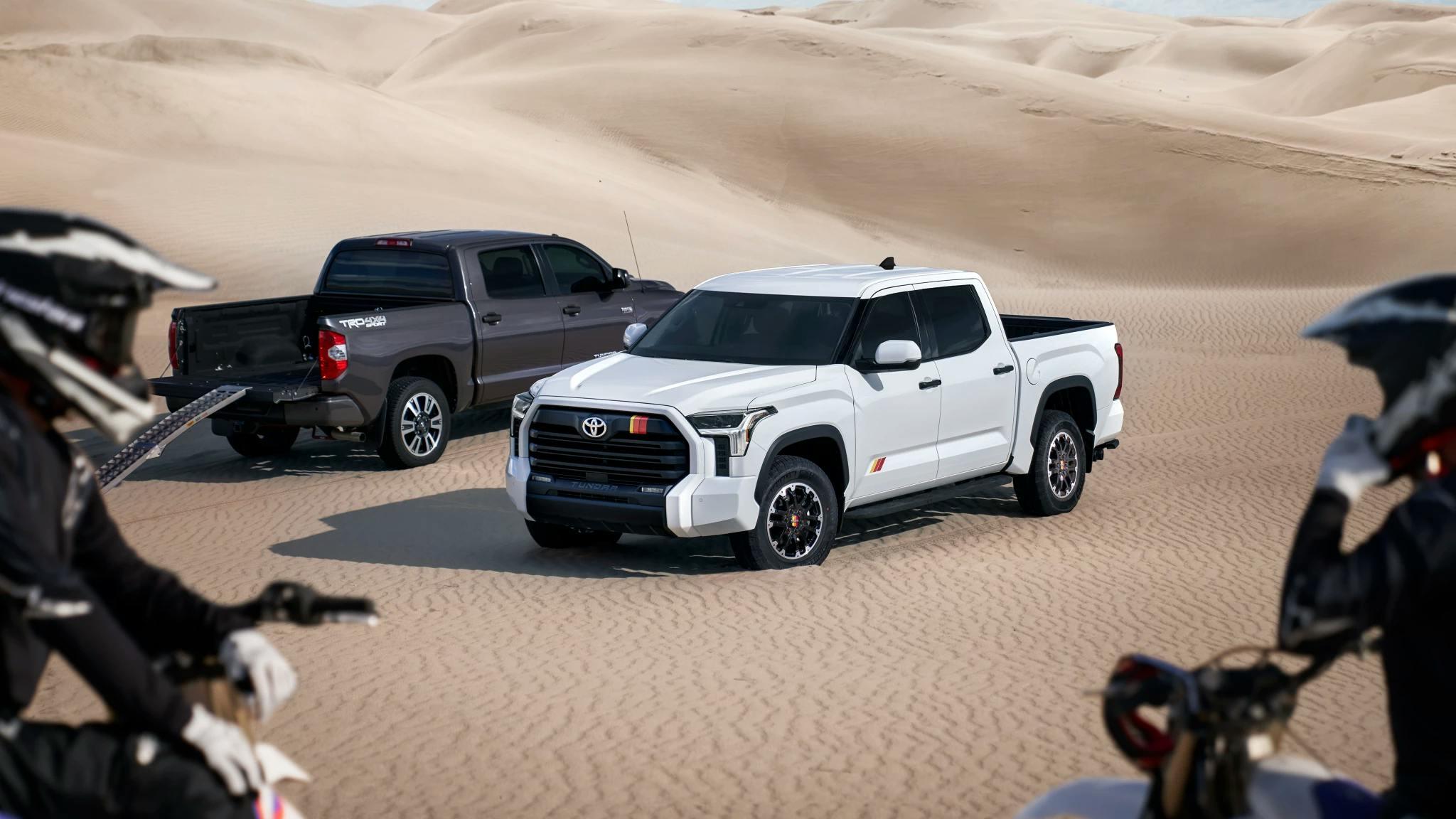 Two Toyota Tundra parked on a sand dune with two motor bike riders in the foreground.