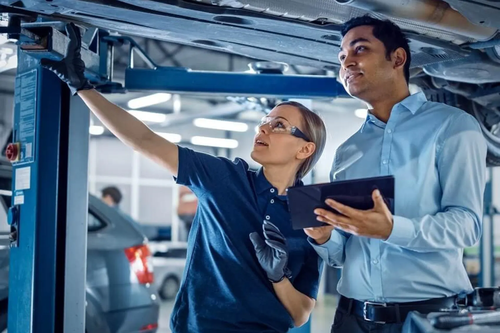 A female mechanic points to the underebelly of a vehicle as a male mechanic holds a tablet