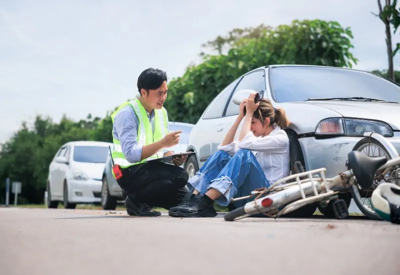 Roadside assistance worker speaking with a distressed woman seated beside a damaged car and fallen motorcycle after an accident.