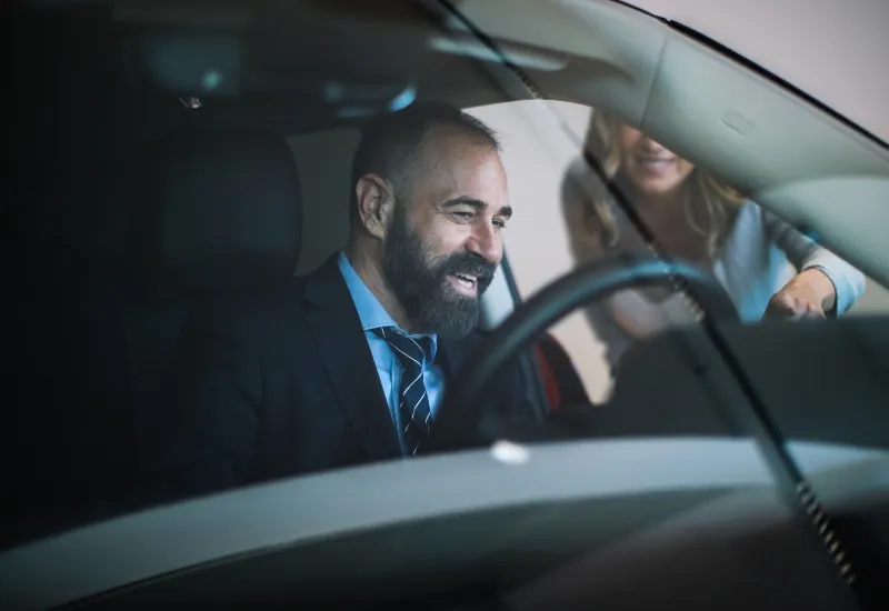 Smiling bearded man seated in the driver’s seat while a woman leans into the car through the open door.