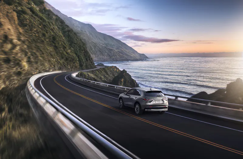 Silver SUV driving along a winding coastal highway beside steep mountains and the ocean at sunset.