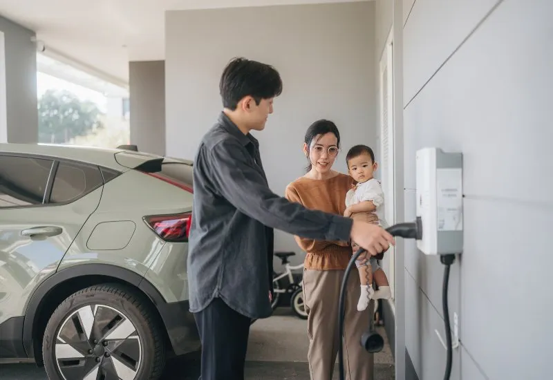 Man plugging an electric vehicle into a home charging station while a woman holds a baby beside the parked SUV.