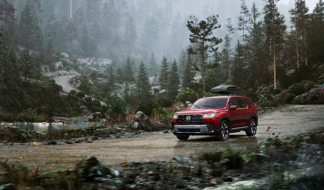 Red Honda SUV with a roof cargo box driving through a shallow stream on a misty mountain road surrounded by pine forest.