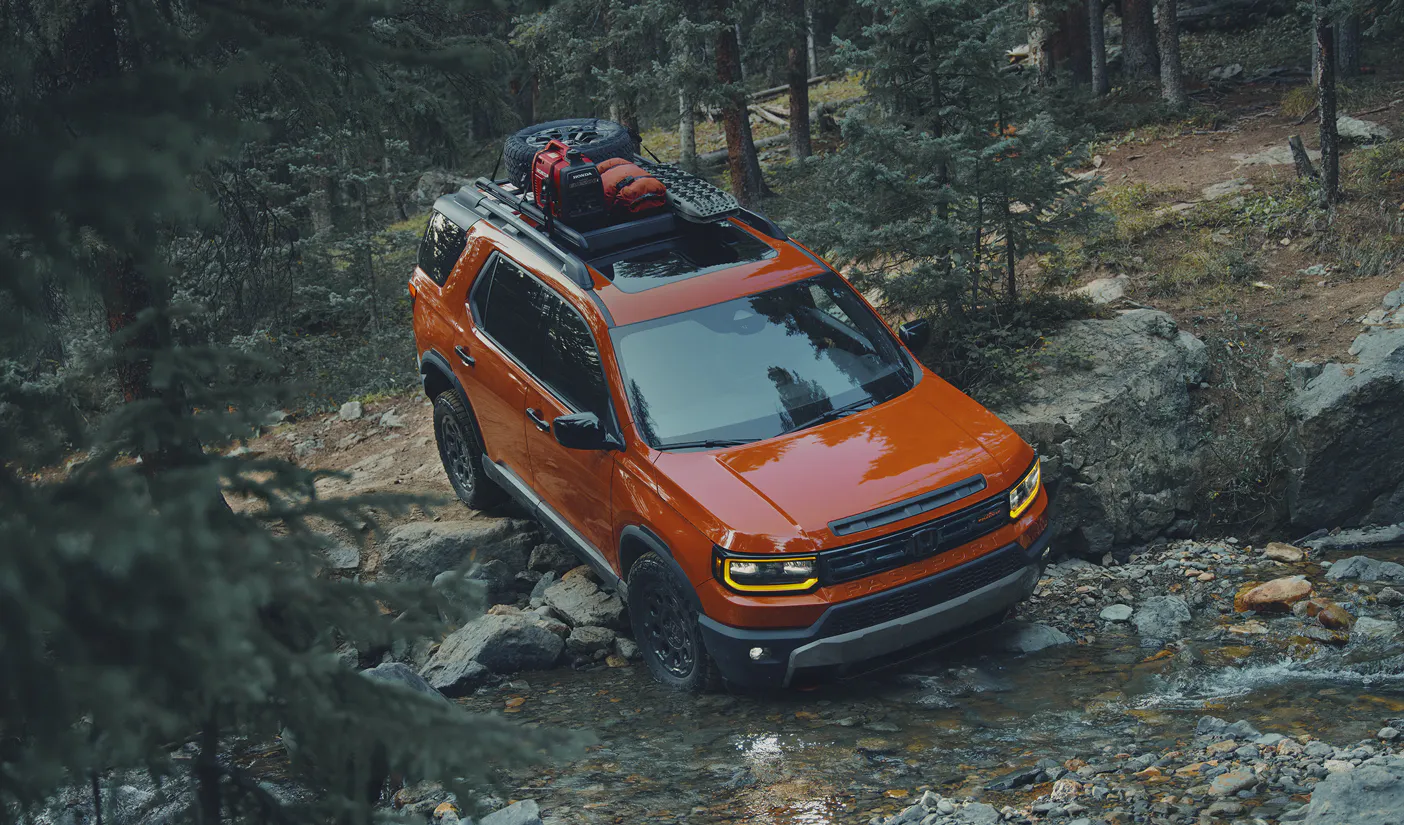 Orange off-road Honda SUV with roof-mounted gear navigating a rocky stream through a dense mountain forest.