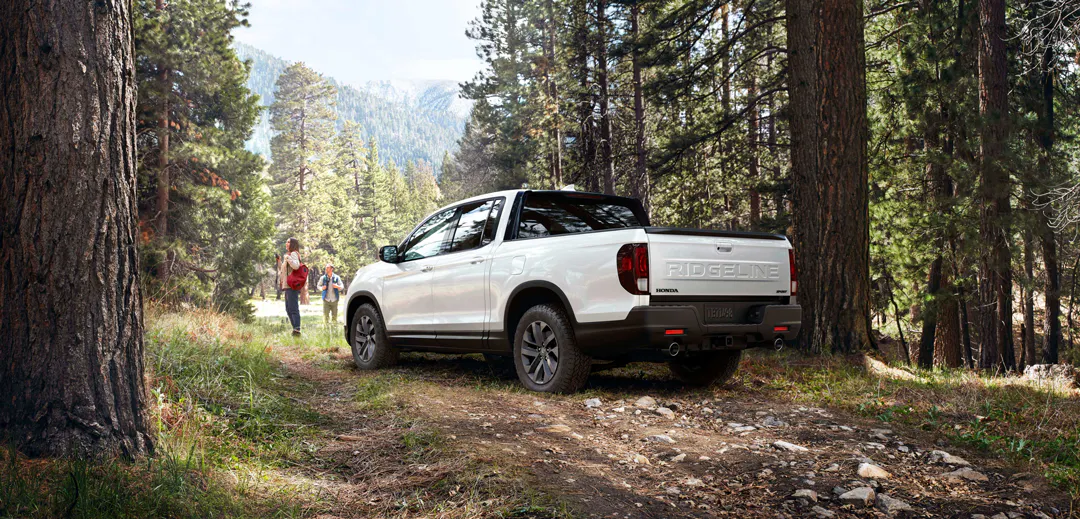 White Honda Ridgeline pickup truck parked on a rocky forest trail while hikers walk nearby among tall pine trees and distant mountains.