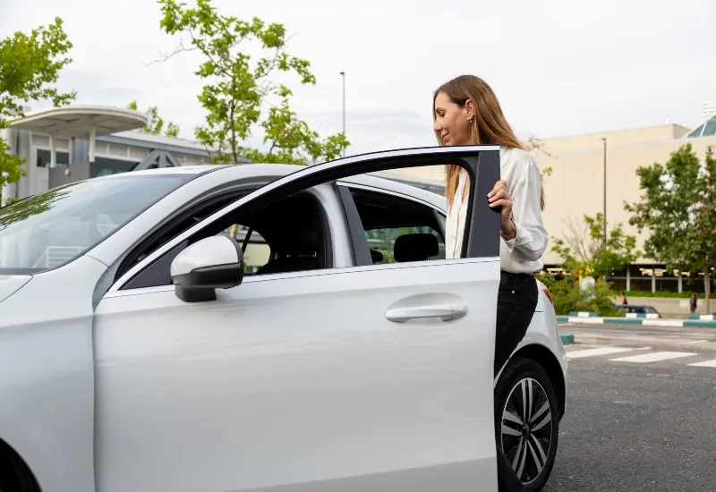 Woman standing beside a white car with the driver’s door open in an outdoor parking area.