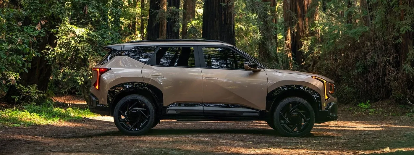 Tan Kia Sportage SUV parked in a lush redwood forest, showcasing its rugged design amid towering trees and natural surroundings.