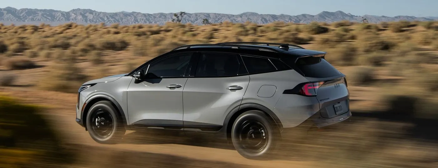 Silver Kia Sportage SUV driving through a desert landscape with mountains in the distance, kicking up dust on an unpaved road.