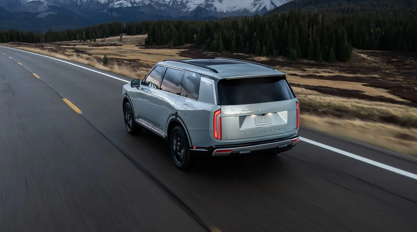 Silver Kia Telluride SUV driving on a scenic highway with evergreen forests and snow-capped mountains in the background.