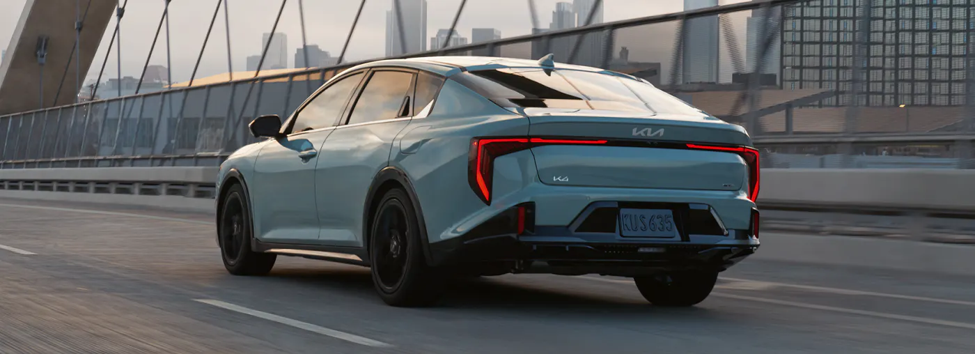 Light blue Kia K4 sedan driving across a modern city bridge with the downtown skyline in the background at sunset.