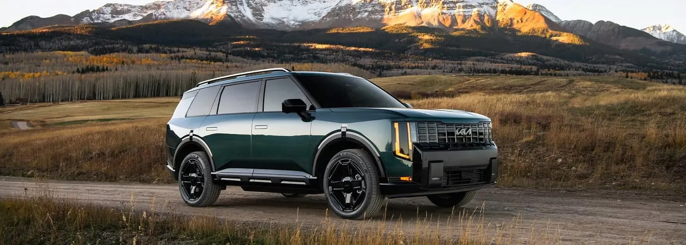 Dark green Kia Telluride SUV parked on a rural road with snow-capped mountains and golden grasslands in the background at sunset.