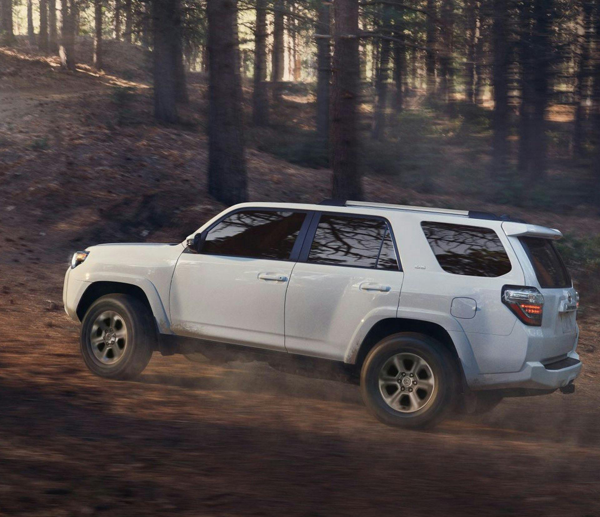 A white Toyota 4Runner drives through a dirt road in the woods with trees in the background