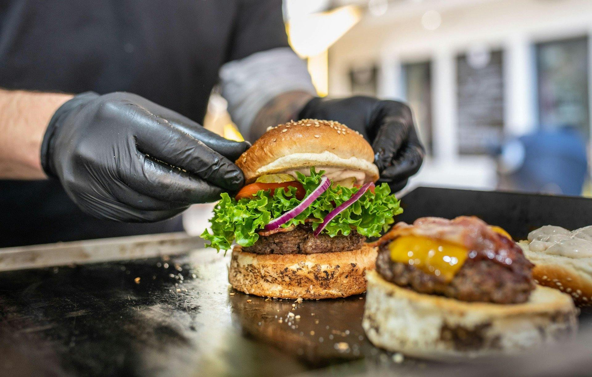 A Chef places a sesame seed bum on top of a large hamburger on the griddle