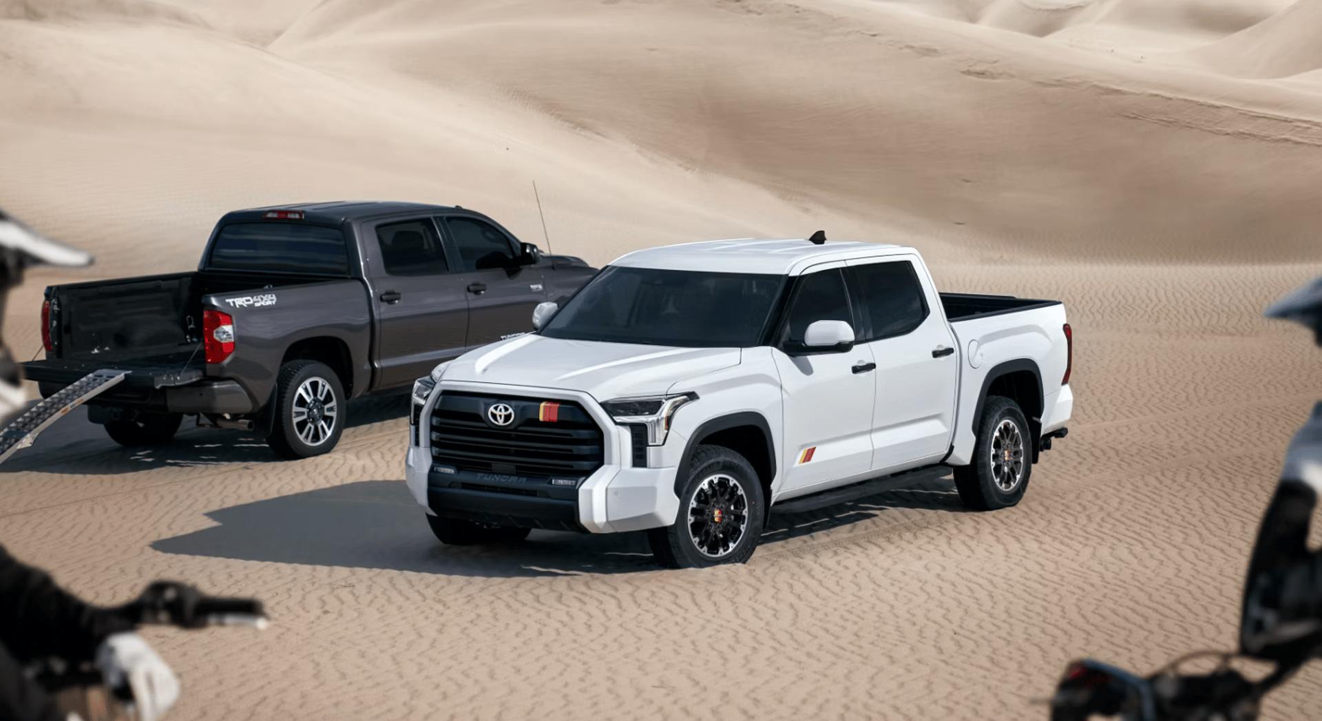 Two Toyota Tundra trucks parked in the sand with two dirt bikes overlooking them.